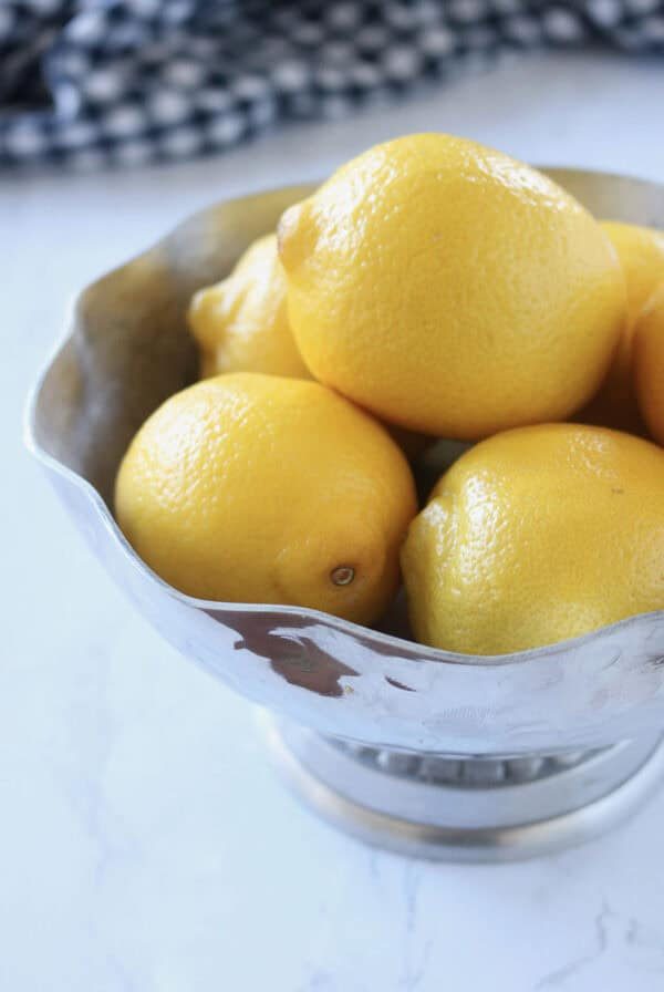 A silver bowl filled with several whole yellow lemons sits on a white marble surface, with a blurred black and white checkered cloth in the background.