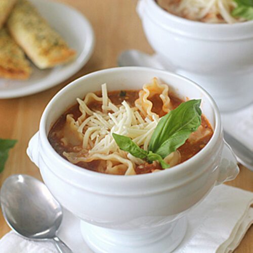 A white bowl filled with lasagna soup, topped with shredded cheese and fresh basil, sits on a napkin with a spoon beside it. Breadsticks and basil leaves are visible in the background.