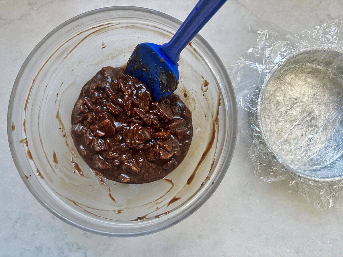 A glass bowl with chocolate and nuts mixture is being stirred with a blue spatula. Next to it is a smaller bowl lined with plastic wrap on a white countertop.