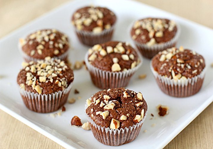 Seven chocolate muffins topped with chopped nuts are arranged on a white square plate, with a few nut pieces scattered around the muffins.