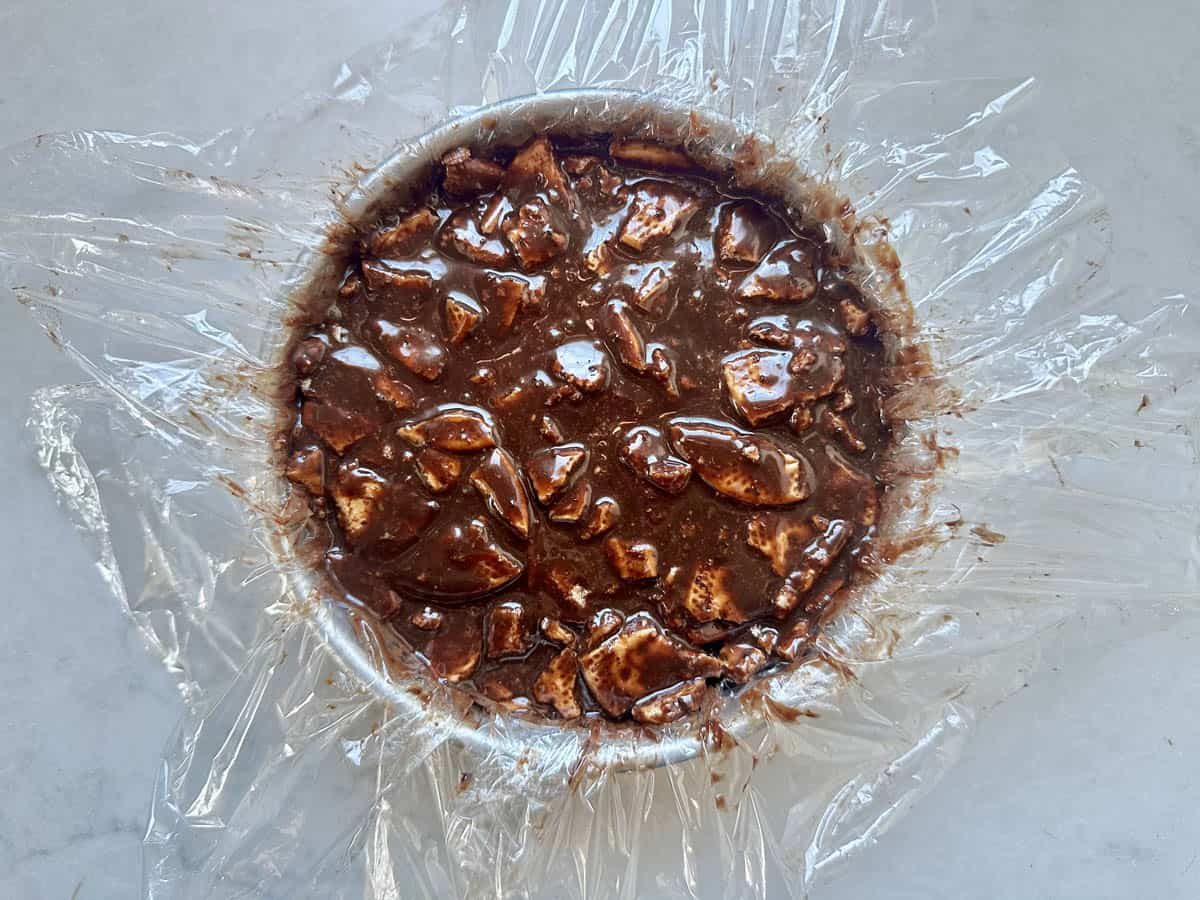 A round dish lined with plastic wrap is filled with a chocolate mixture containing visible chunks, sitting on a light-colored surface.