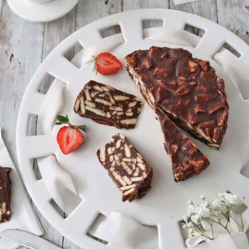 A white plate holds slices of chocolate biscuit cake, surrounded by fresh strawberries. A bowl of strawberries is nearby, and a knife rests on the table. The scene is set on a rustic wooden surface.