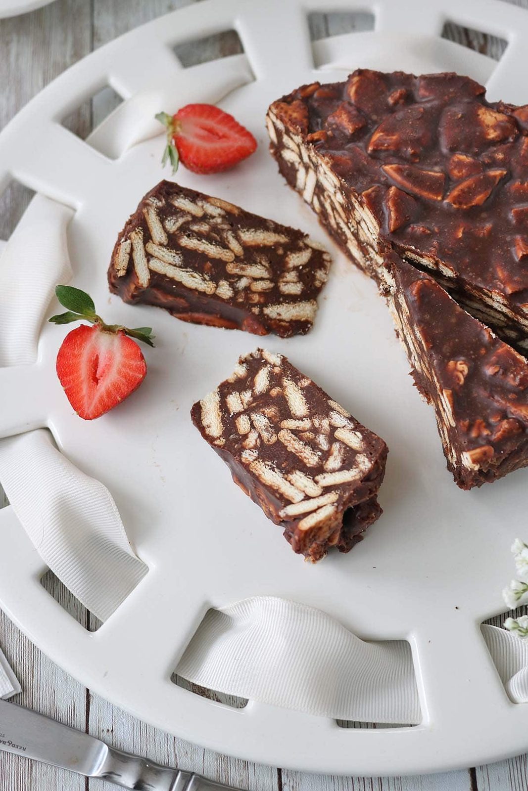A white plate with slices of chocolate biscuit cake, garnished with halved strawberries. The cake shows a marbled pattern of biscuits and chocolate. A white cloth napkin is partially visible on the plate.