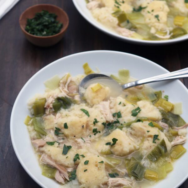 Two white bowls filled with chicken and dumplings soup, featuring chunks of chicken, dumplings, celery, and broth, garnished with chopped herbs. Spoons rest in the bowls, with a small bowl of herbs beside them.