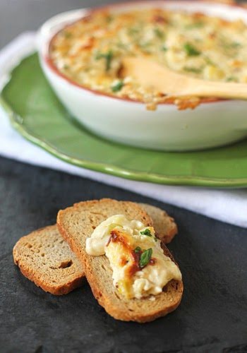 Two slices of toasted bread, one topped with creamy, cheesy dip and herbs, sit on a dark surface. In the background, a baking dish filled with hot dip rests on a green plate.