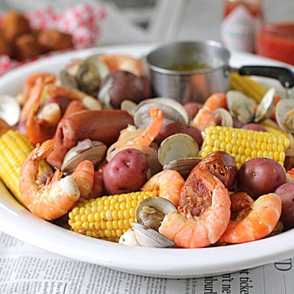 A white platter filled with shrimp, clams, sausage, corn on the cob, and red potatoes, with a small cup of melted butter; newspaper and cocktail sauce are visible in the background.