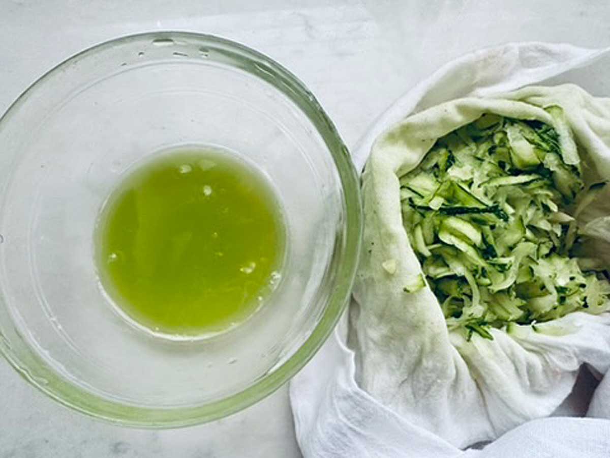 A glass bowl with green liquid sits next to grated zucchini wrapped in a white cloth, likely to squeeze out excess moisture from the zucchini.