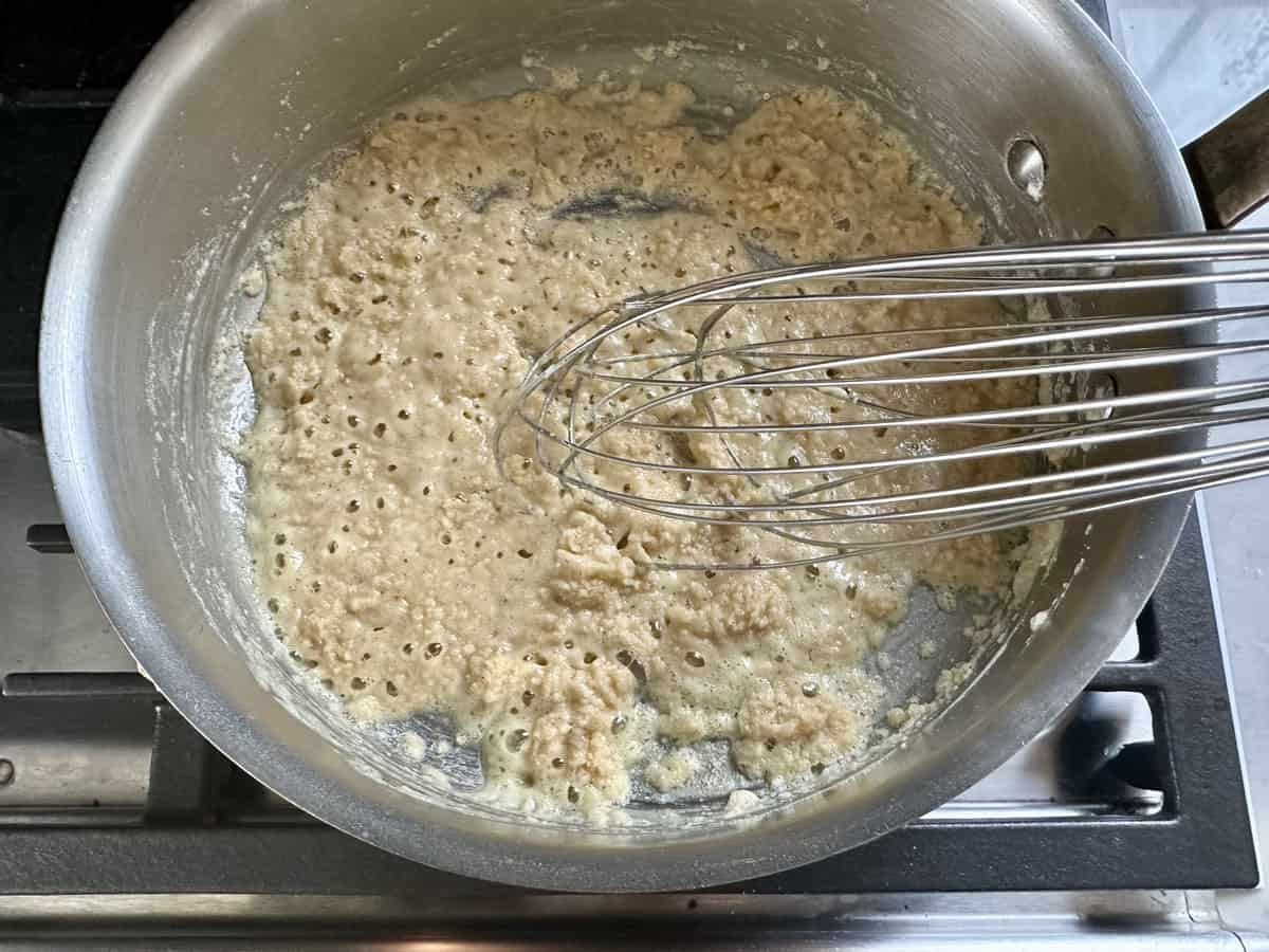 A metal whisk stirring a bubbling mixture of flour and fat in a stainless steel pan on a stovetop, making a light brown roux.