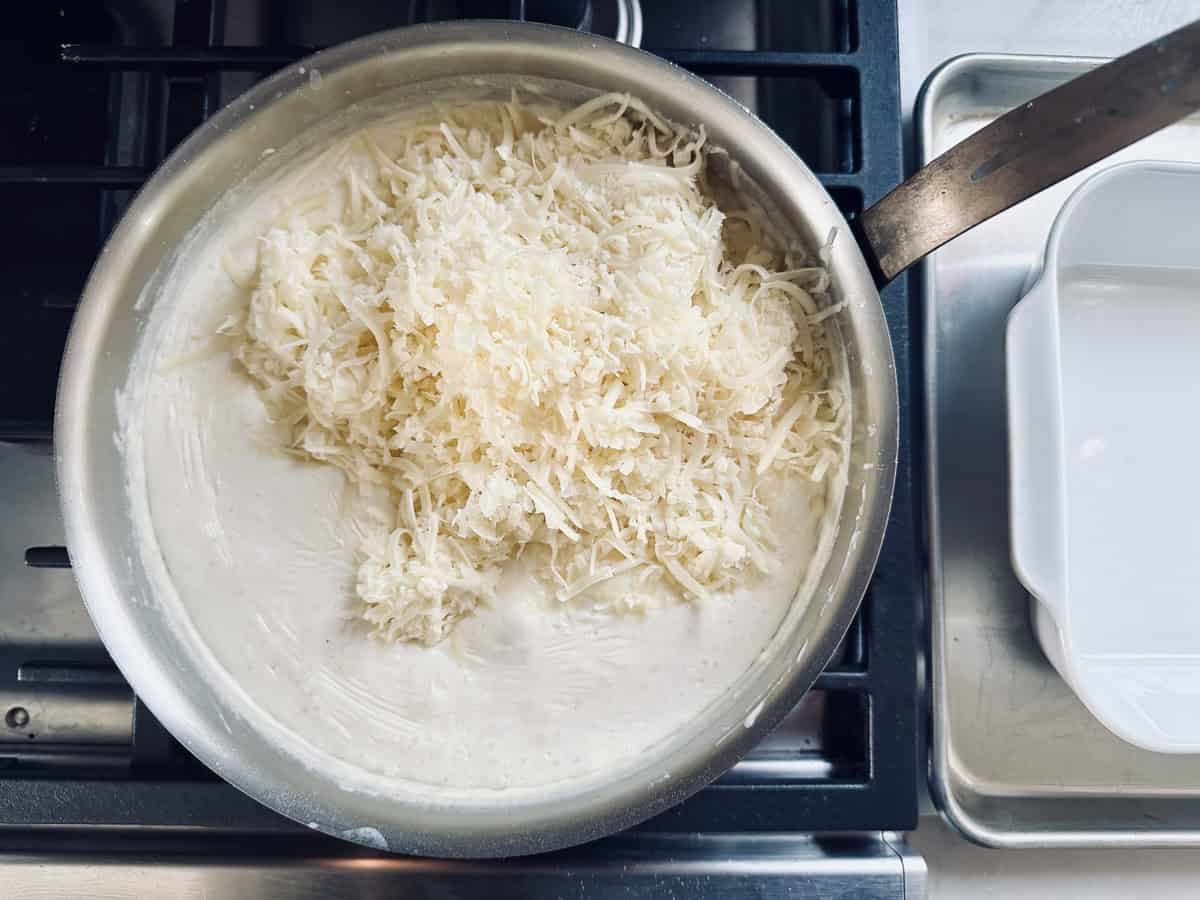 A metal pan on a stove contains a creamy white sauce topped with a generous pile of shredded cheese, ready to be mixed in. A baking dish sits nearby on a metal tray.