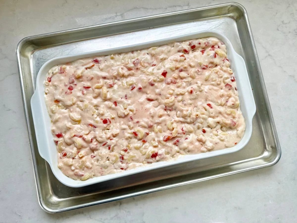 A white baking dish filled with an unbaked creamy mixture containing chopped red and white ingredients, placed on a silver baking tray atop a light-colored countertop.