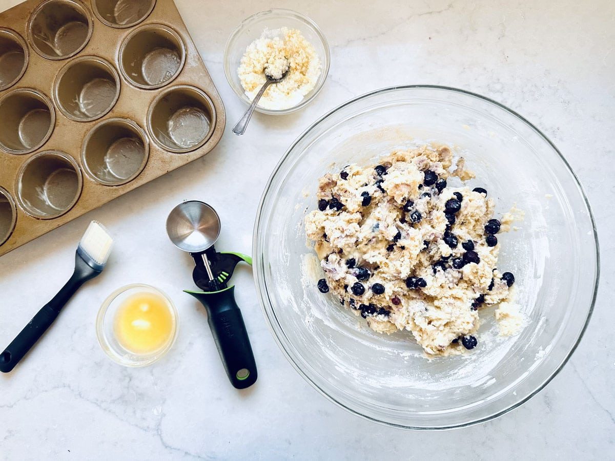 A glass bowl of blueberry muffin batter sits on a white countertop surrounded by a muffin tin, a pastry brush, a bowl of beaten egg, a cookie scoop, and a small bowl of crumb topping with a spoon.