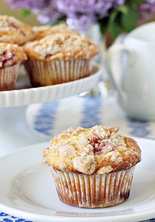 A crumb-topped muffin sits on a white plate, with more muffins displayed on a cake stand in the background. A teapot and blurred flowers are also visible.