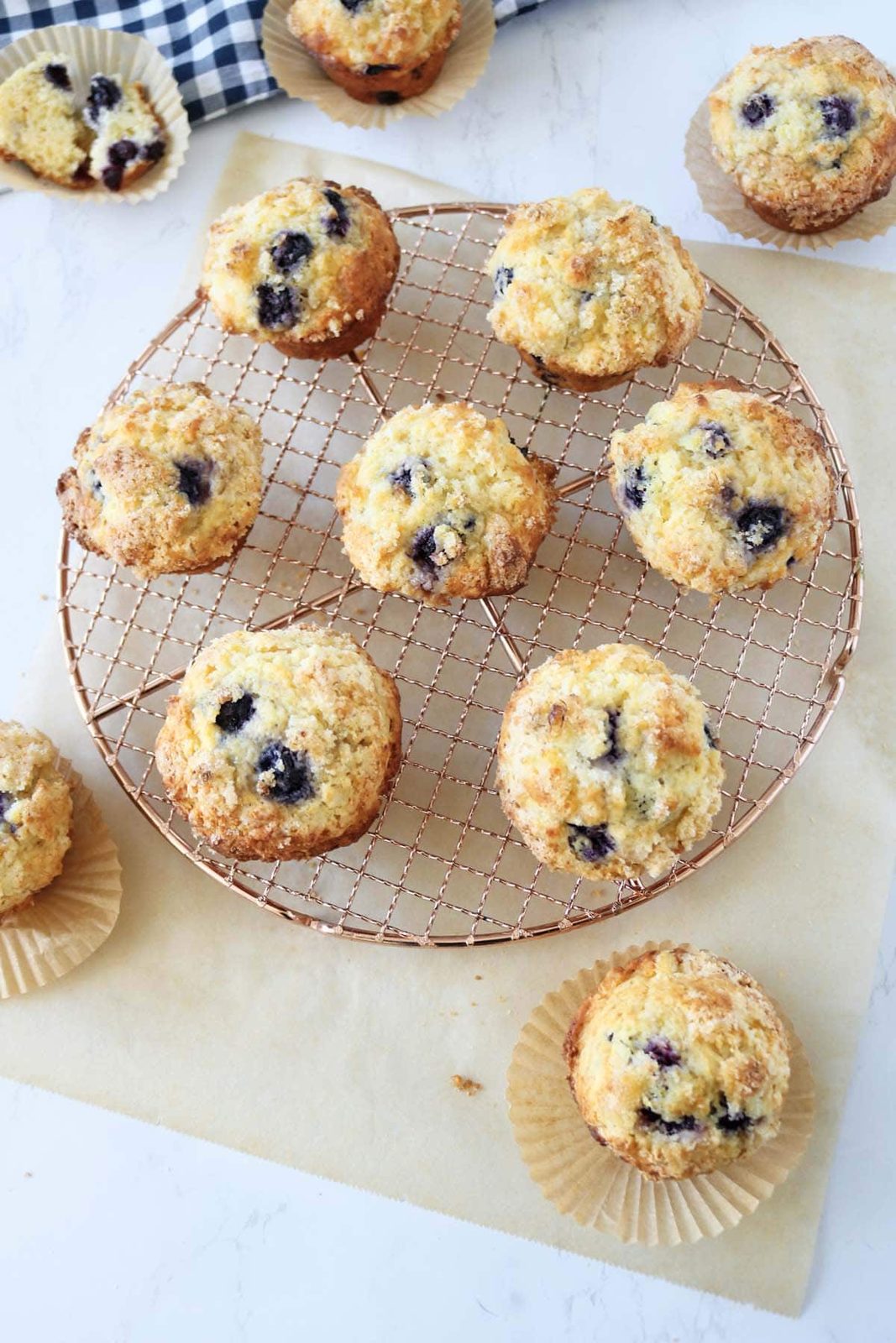 Nine blueberry muffins on a round cooling rack, with a few more muffins on parchment paper nearby. The muffins are golden brown with visible blueberries and a sugar topping. A checkered cloth is partially visible in the corner.