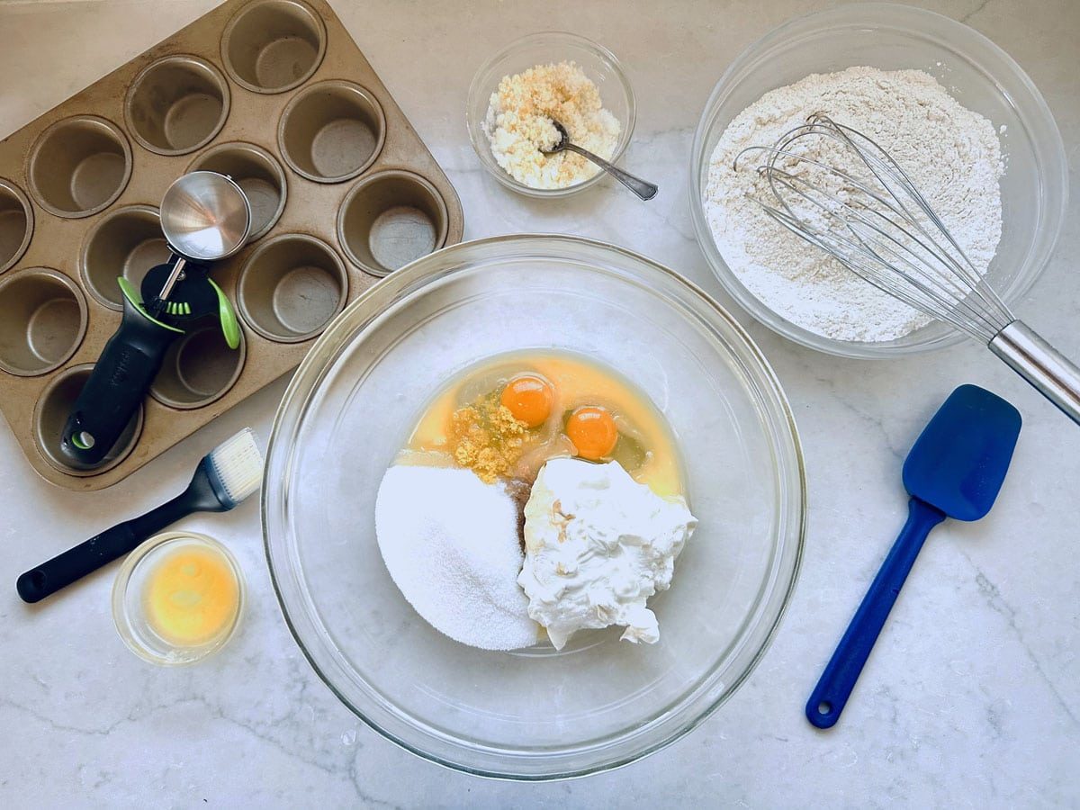 A muffin tin, ice cream scoop, brush, bowl of flour, bowl with eggs, sugar, and sour cream, small bowls of cheese and egg wash, and a spatula are arranged on a white countertop for baking.