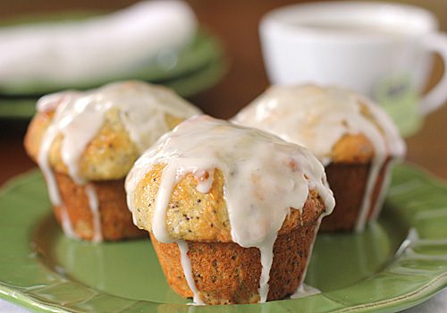 Three poppy seed muffins topped with white icing glaze sit on a green plate, with a cup and saucer blurred in the background.