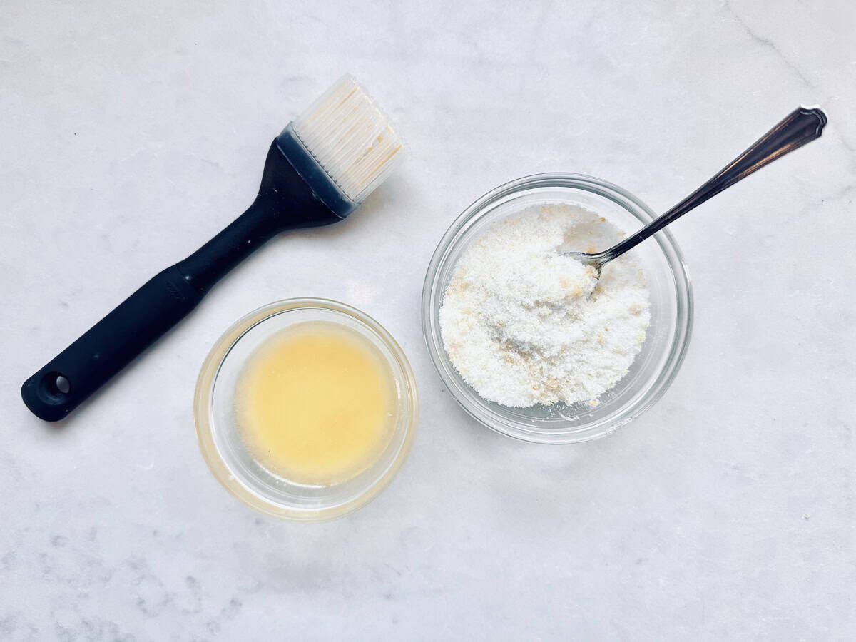 A black pastry brush, a small bowl of melted butter, and a small bowl of grated Parmesan cheese with a spoon, all arranged on a white marble surface.