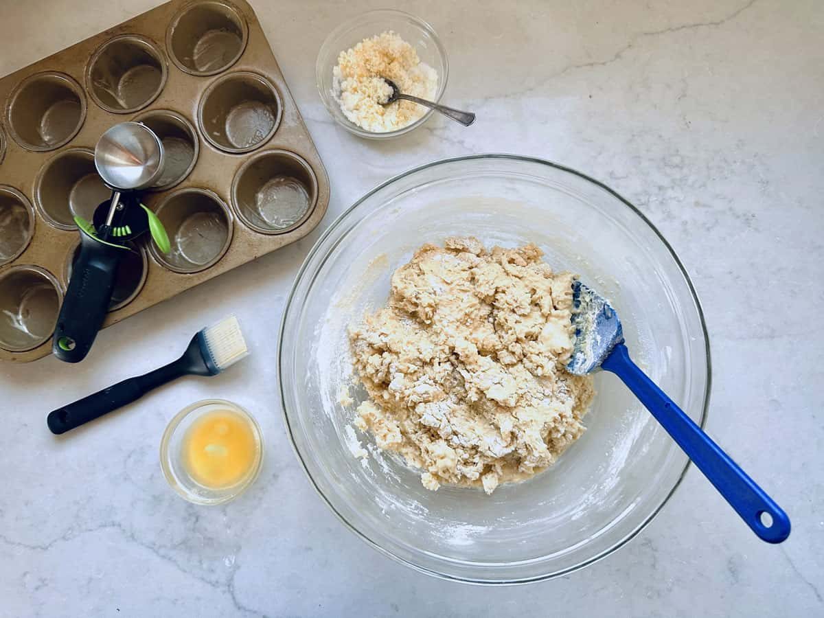 A glass bowl with biscuit dough and a blue spatula sits on a countertop, surrounded by a muffin tin, measuring tools, a pastry brush, a small bowl of egg wash, and a bowl of crumbled cheese.