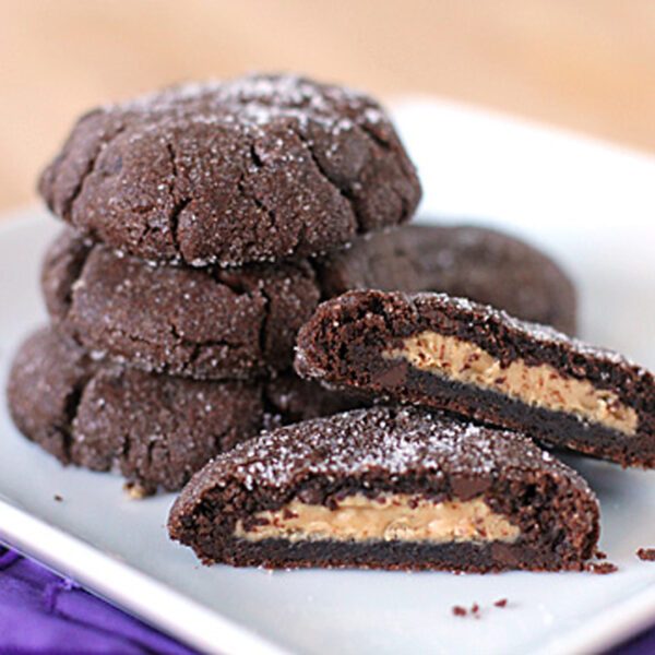 A plate of chocolate cookies stacked and arranged on a white square dish, with one cookie cut open to reveal a creamy peanut butter filling inside. Sugar is sprinkled on top of the cookies.