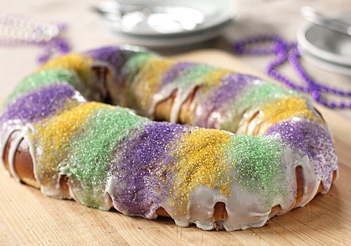 A ring-shaped King Cake covered in white icing and decorated with green, purple, and yellow sugar, sitting on a wooden surface with blurred plates and beads in the background.