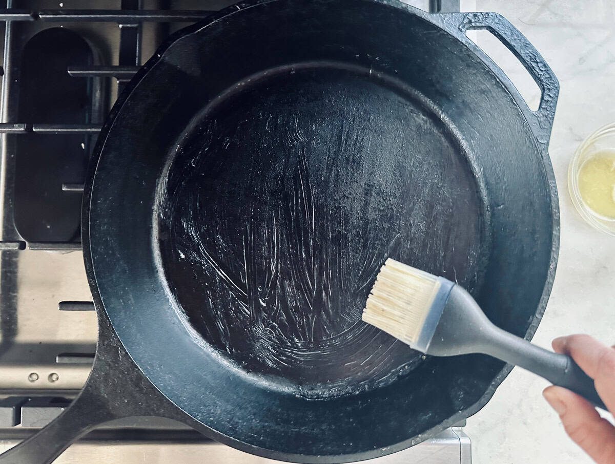 A hand uses a silicone brush to spread oil or butter onto a black cast iron skillet on a stovetop. A small bowl with liquid is visible nearby on a light-colored countertop.