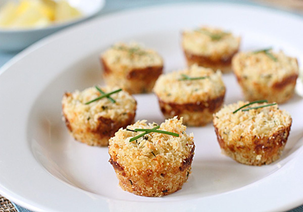 Eight golden-brown, bite-sized appetizers topped with herbs are arranged on a white plate. The food has a crispy texture, and a blurred dish is visible in the background.