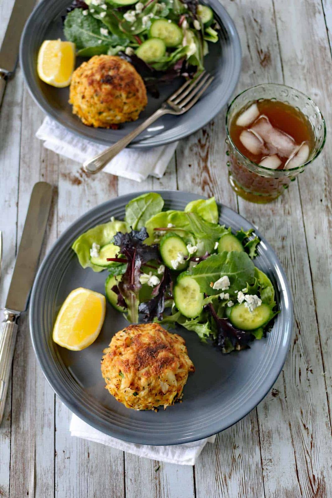 Two gray plates with leafy green salads, cucumber slices, a lemon wedge, and a golden-brown crab cake. A glass of iced tea sits nearby on a rustic wooden table, with silverware and white napkins on the side.