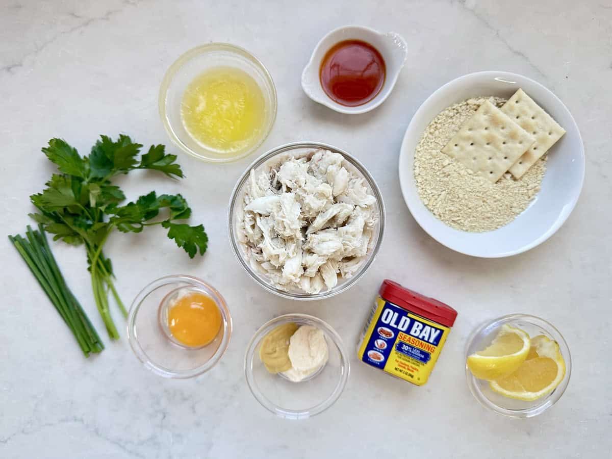 A selection of ingredients for crab cakes: crab meat, egg yolk, melted butter, hot sauce, mayonnaise, mustard, Old Bay seasoning, parsley, chives, lemon wedges, oyster crackers, and saltine crackers on a white surface.