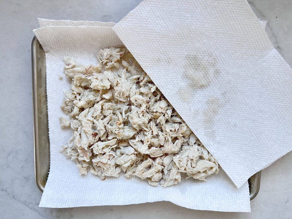 A pile of shredded cooked chicken is spread on a paper towel-lined tray, with another paper towel partially covering it, soaking up moisture. The tray sits on a light-colored surface.