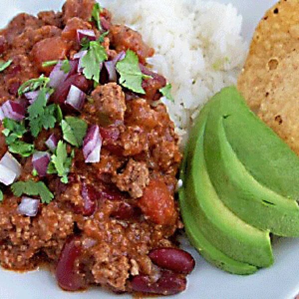 A plate with chili con carne topped with chopped red onion and cilantro, served with white rice, sliced avocado, and a tortilla chip.