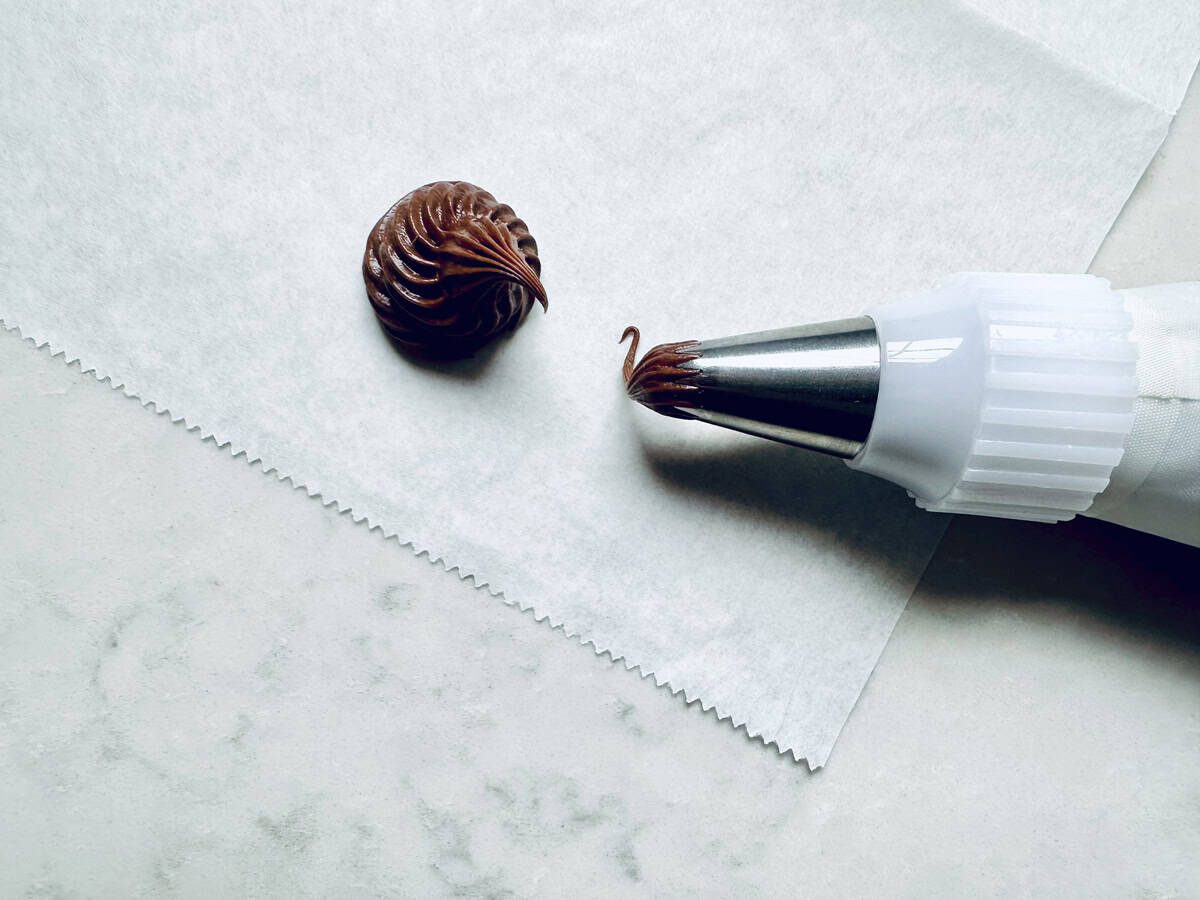 A piping bag with a metal tip is squeezing a swirl of chocolate frosting onto a piece of parchment paper on a light-colored surface.
