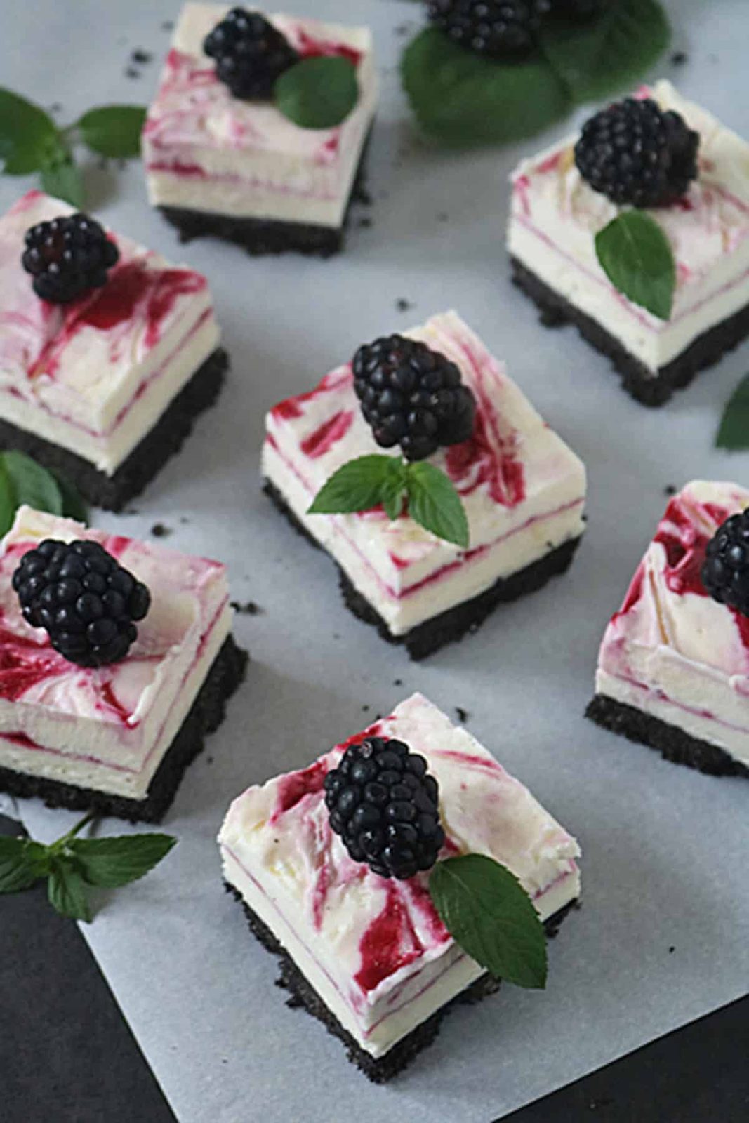 Squares of creamy cheesecake with a chocolate crumb crust, topped with a blackberry swirl, fresh blackberries, and mint leaves, arranged on parchment paper.
