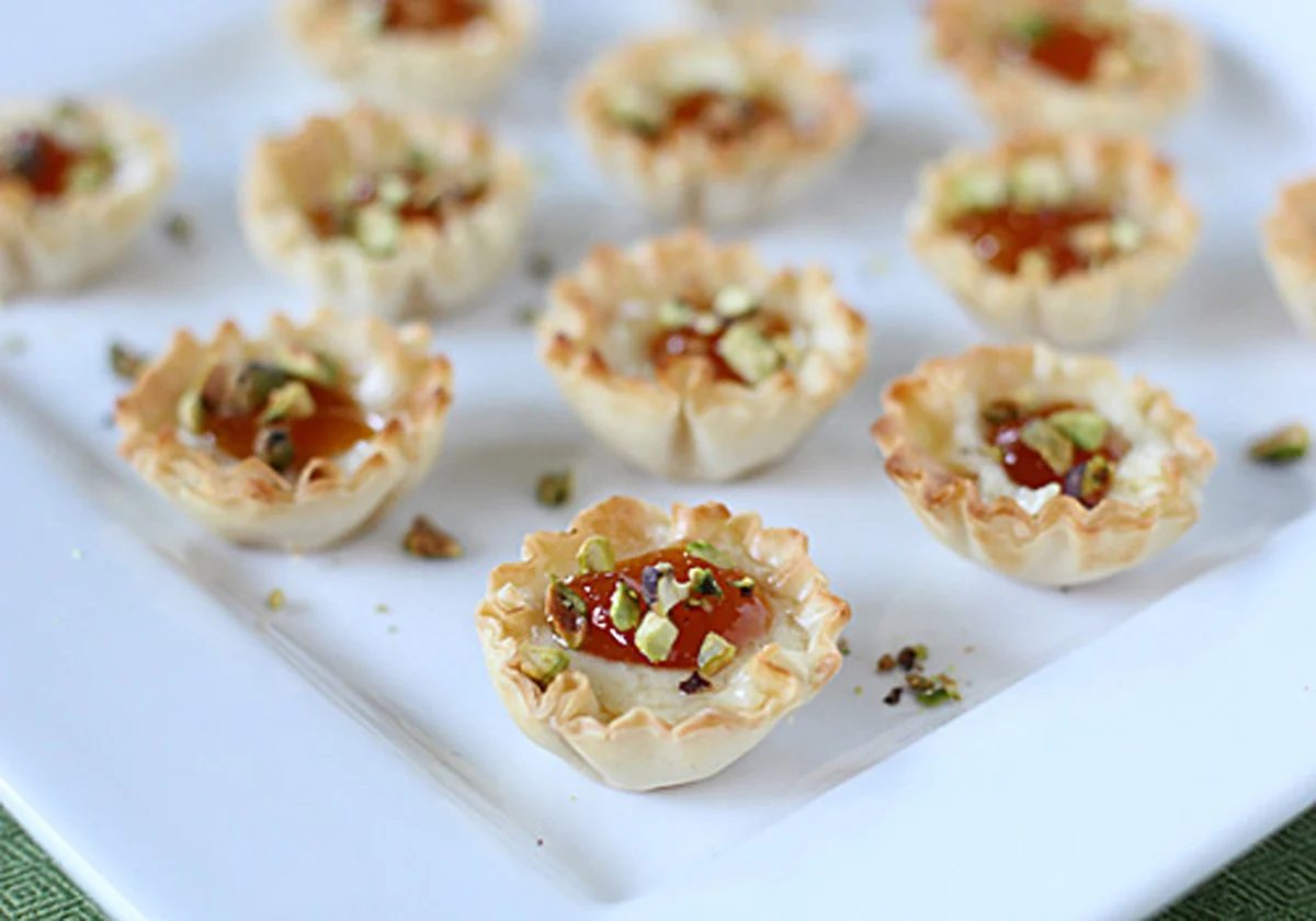 Mini phyllo pastry cups filled with a creamy mixture, topped with a dollop of jam and chopped pistachios, arranged on a white square plate.