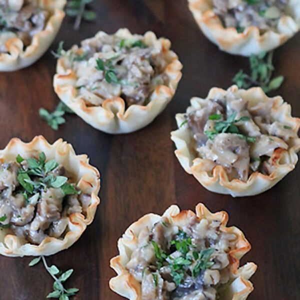 Mini phyllo pastry cups filled with a creamy mushroom mixture, garnished with fresh herbs, are arranged on a dark wooden surface.