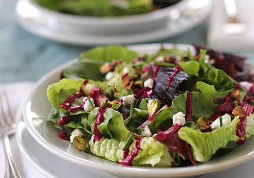 A fresh salad with mixed greens, crumbled cheese, nuts, and a vibrant purple dressing served on a white plate, with a fork and another plate in the background.