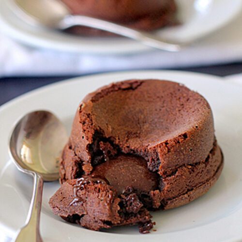 A chocolate lava cake with a gooey center sits on a white plate with a spoon beside it. Another cake and spoon are blurred in the background.