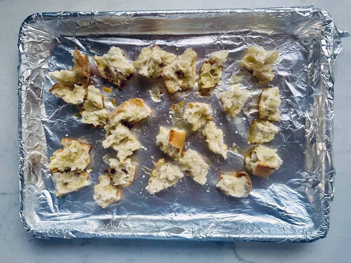 Chunks of torn bread scattered on a foil-lined baking tray, ready for toasting or baking. The bread appears partially crispy and is placed on a light-colored surface.