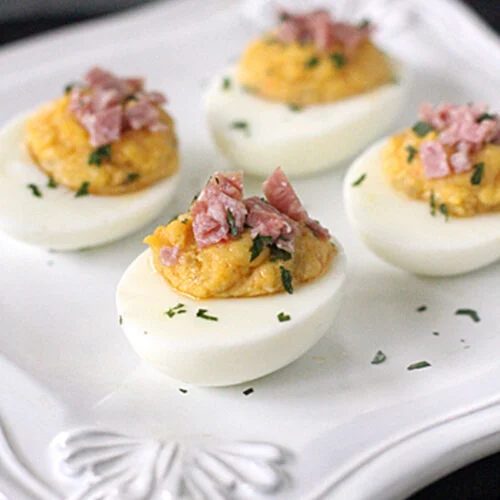 Four deviled eggs topped with chopped ham and sprinkled with herbs are arranged on a white decorative plate.