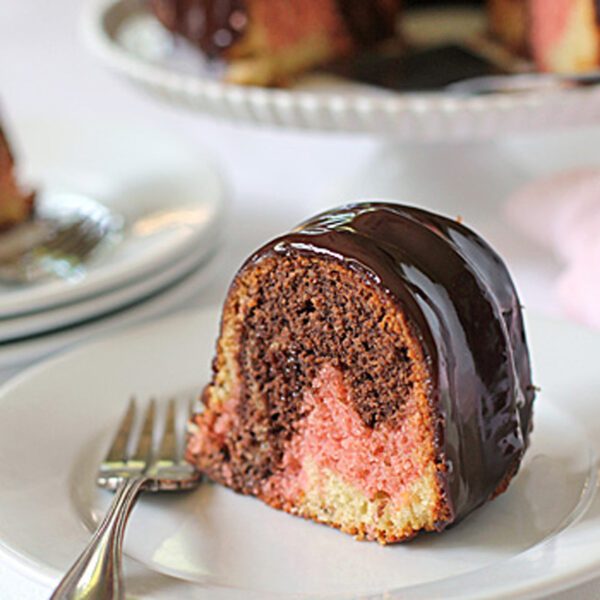 A slice of marble bundt cake with chocolate, vanilla, and pink layers, topped with glossy chocolate glaze, sits on a white plate with a fork beside it. More cake slices are visible in the background.