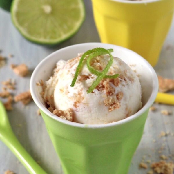 A scoop of ice cream sprinkled with crumbs and topped with lime zest sits in a green cup, with a green spoon nearby. A yellow cup with more ice cream and a halved lime are in the background.