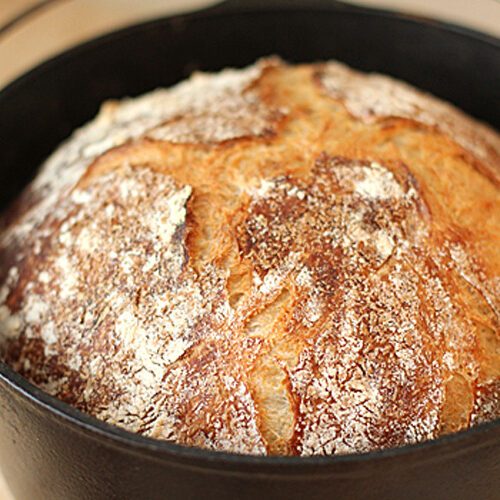 A round, golden-brown loaf of crusty bread with a rustic, cracked surface sits in a black cast iron pot.