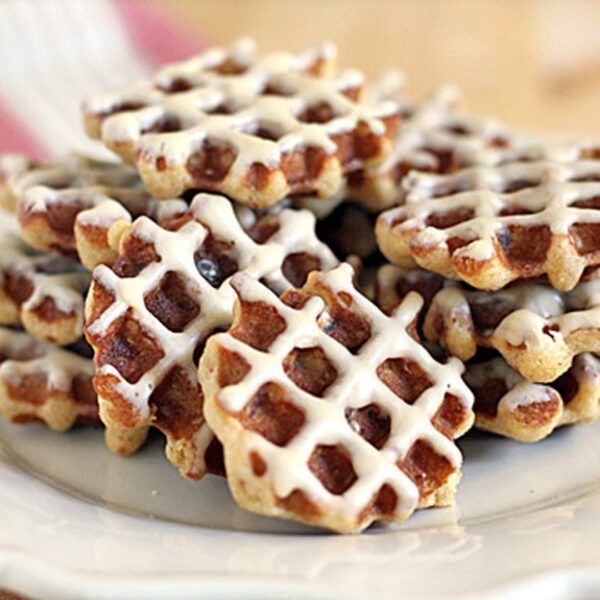 A stack of small waffle cookies drizzled with white icing, arranged on a white plate with a fork in the background.
