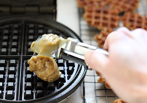A hand uses a cookie scoop to drop cookie dough onto a waffle maker, with baked waffle cookies cooling on a rack in the background.