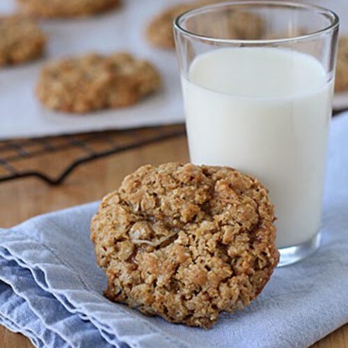 A glass of milk and an oatmeal cookie rest on a folded blue cloth. More oatmeal cookies are visible in the background, slightly out of focus, on a wire rack and baking sheet.