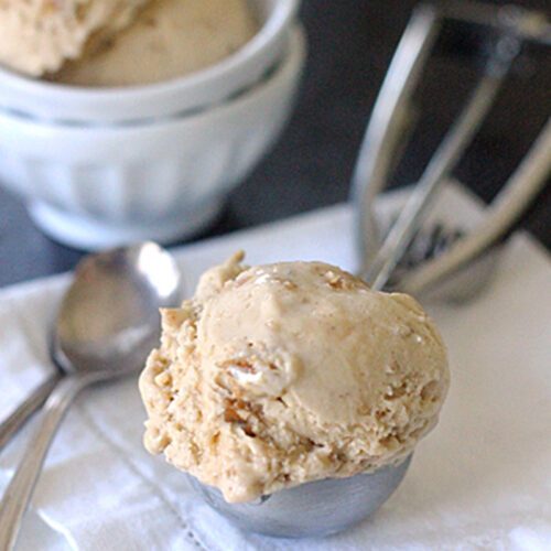 A scoop of light brown ice cream in a metal scoop rests on a white cloth next to two silver spoons, with a bowl containing more ice cream and an ice cream scooper in the background.