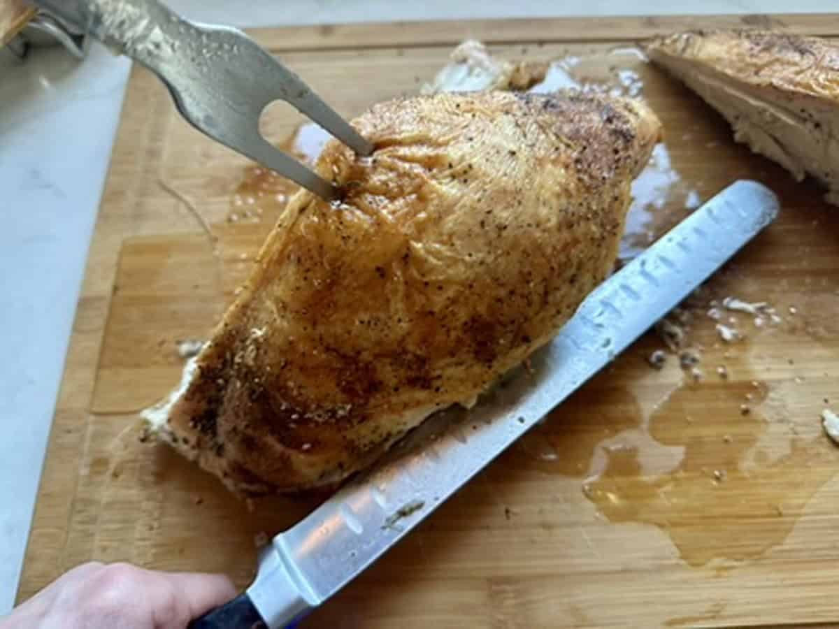 A hand holding a knife slices into a seasoned, cooked chicken breast held steady by a fork on a wooden cutting board.