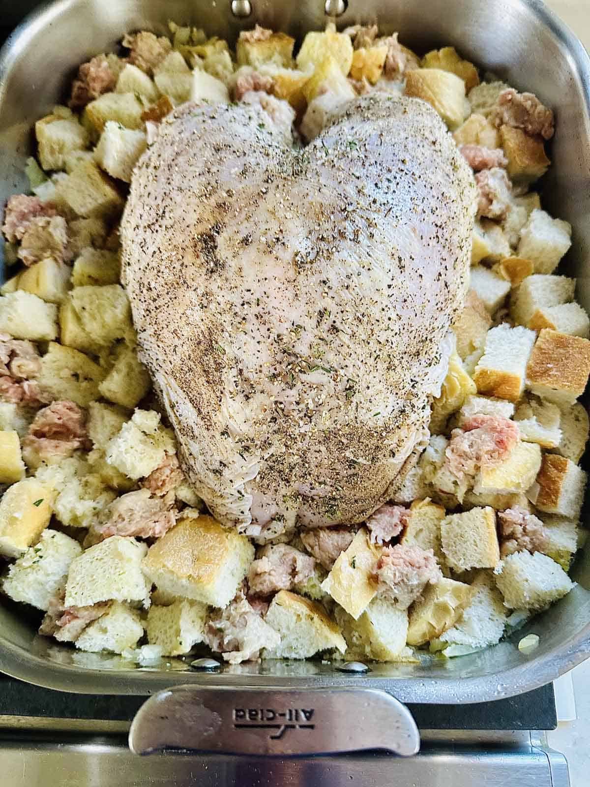 A seasoned turkey breast sits on top of cubed bread and sausage stuffing in a roasting pan, ready to be cooked.