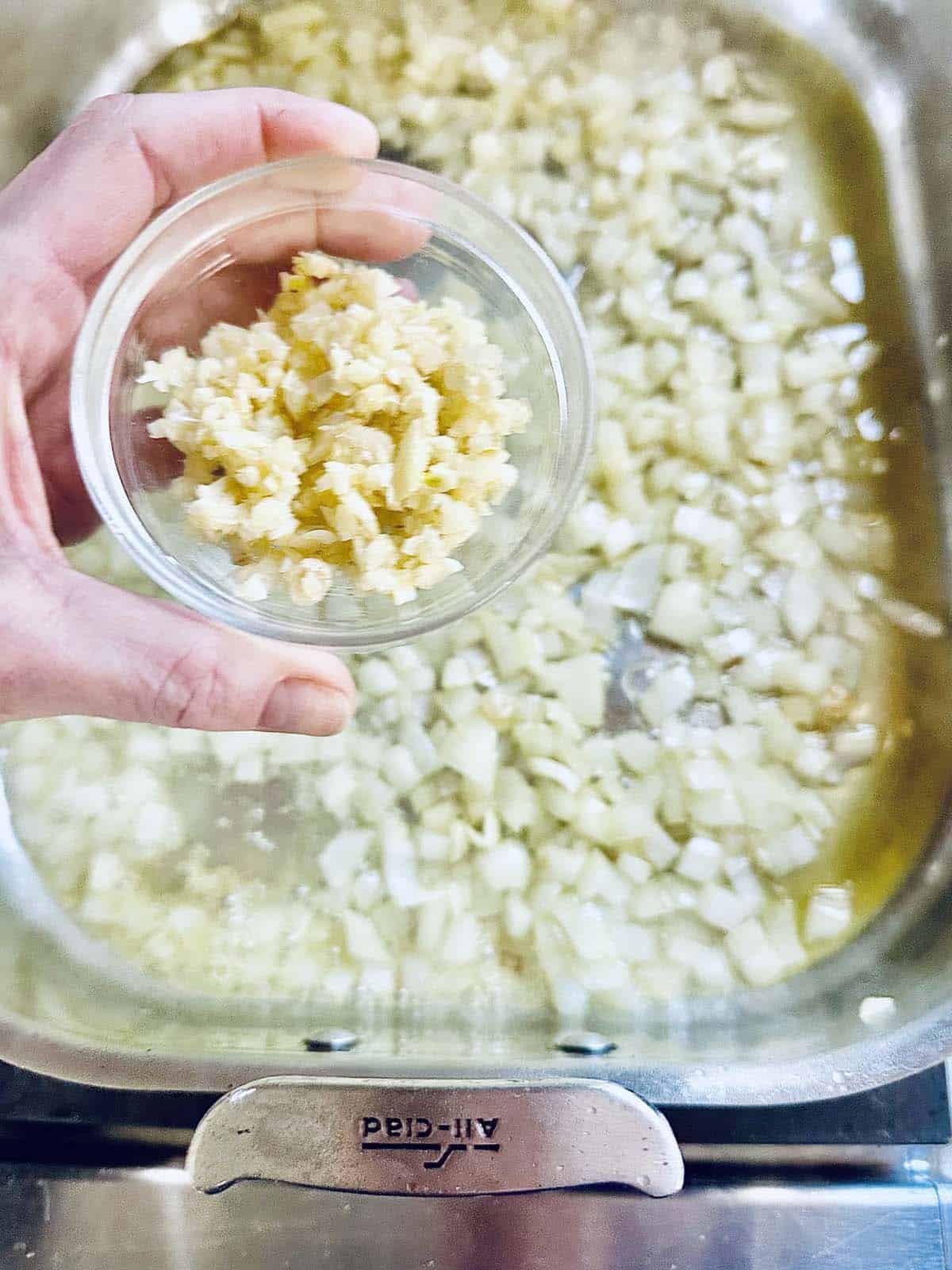 A hand holds a small bowl of chopped garlic above a metal pan filled with diced onions cooking in oil.