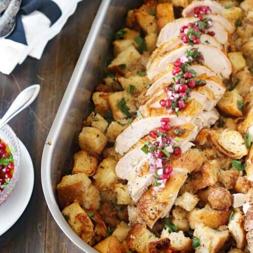A roasting pan filled with cubed bread stuffing, topped with sliced roasted turkey breast garnished with herbs and pomegranate seeds. A spoon and napkins are on the side.