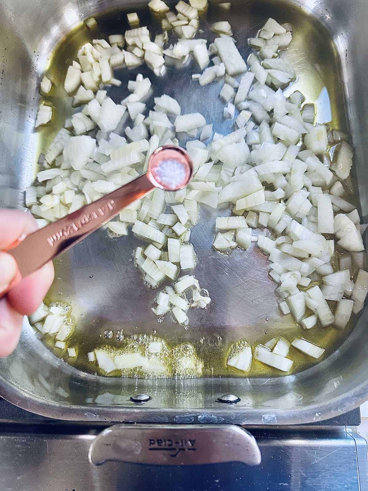 A hand holds a measuring spoon of salt above a pan with chopped onions sautéing in oil on a stovetop.
