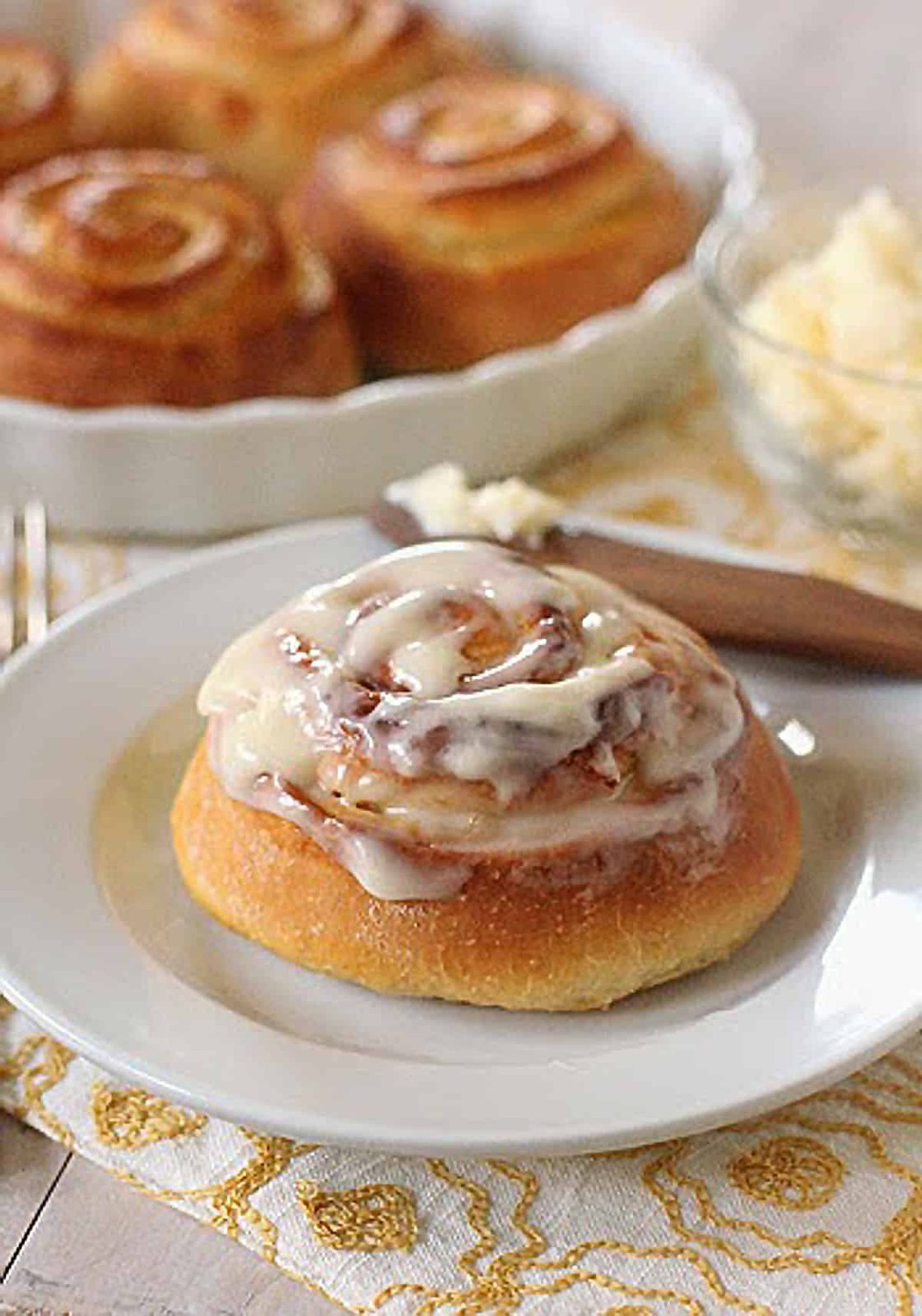A frosted cinnamon roll sits on a white plate, with more cinnamon rolls on a dish and a bowl of butter in the background on a patterned yellow and white cloth.